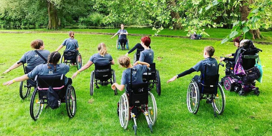 A group of 9 wheelchair users dancers on a grassed area. They are facing away from the camera towards the instructor with their arms to their side.