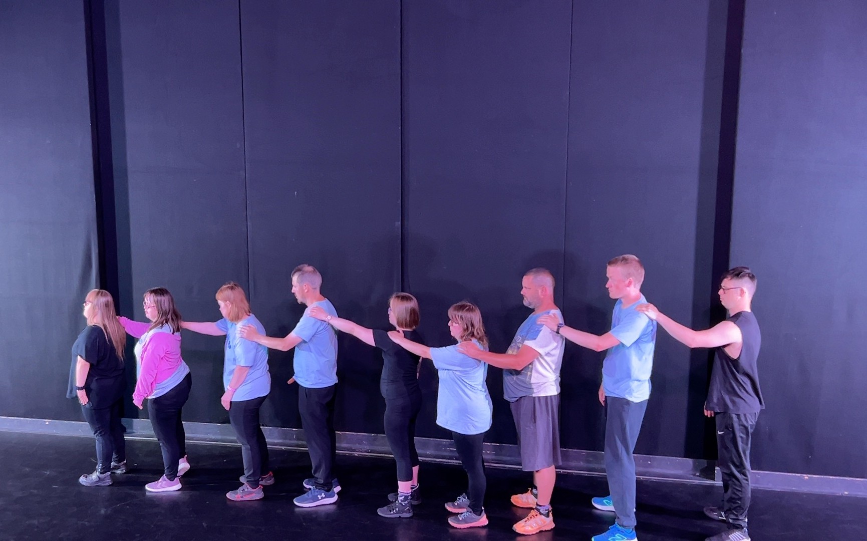 A group of dancers, all standing together in a line, holding each others shoulders.