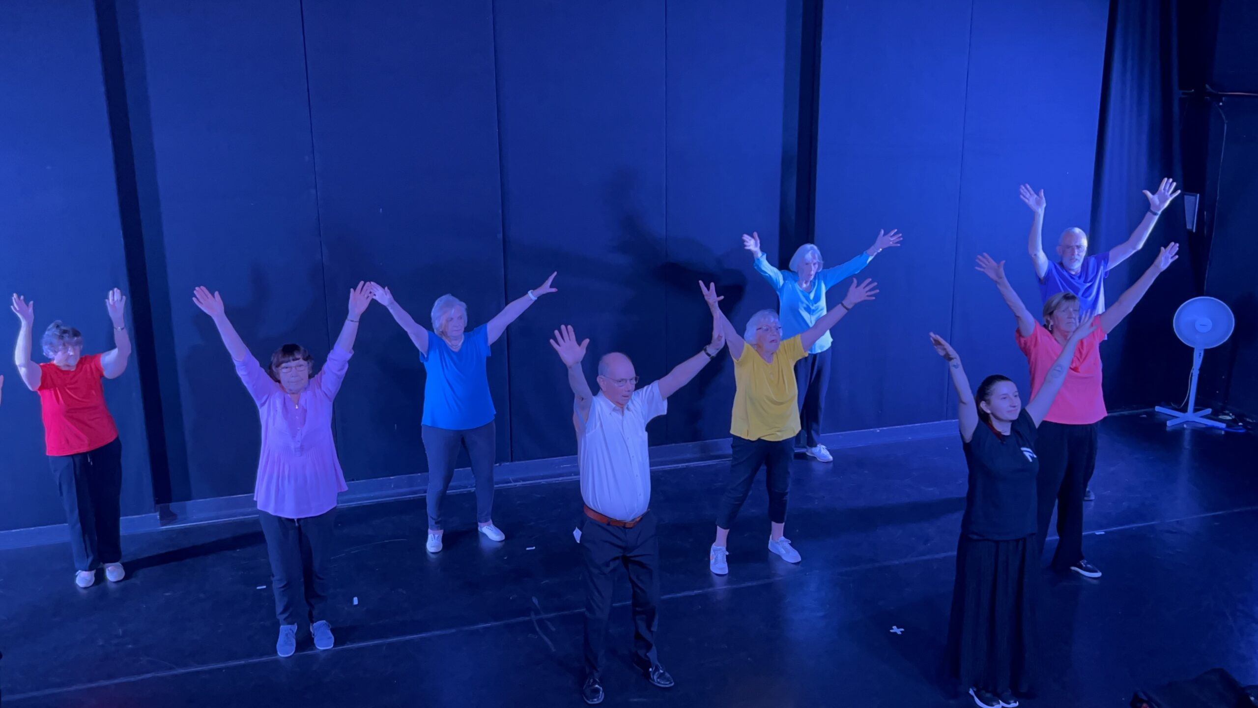 A group of older dancers and their teacher all dancing together with colourful tops, and their arms in the air in a "V" poae