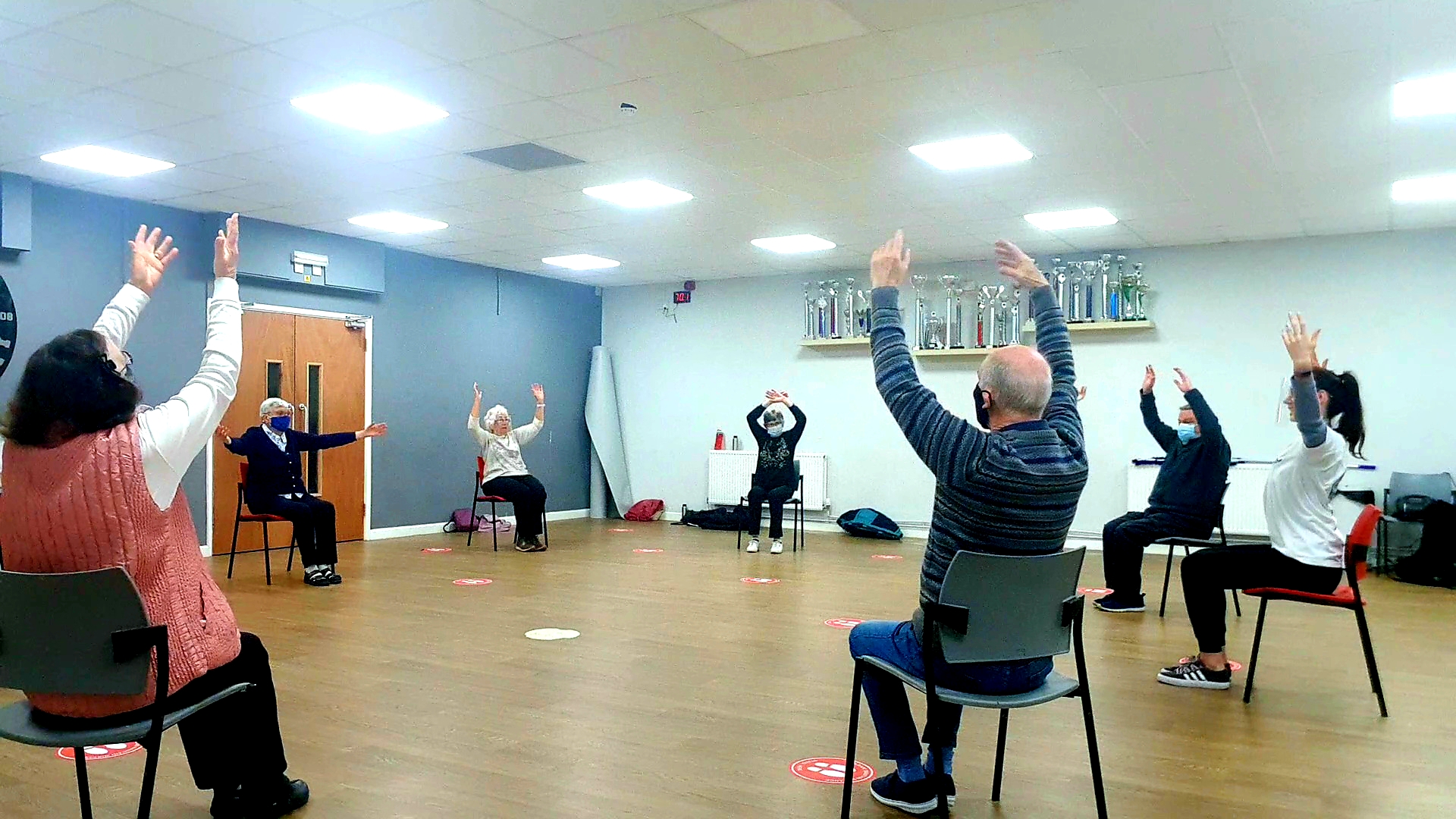 A group of older dancers all sat down, in a circle, with their arms in the air.